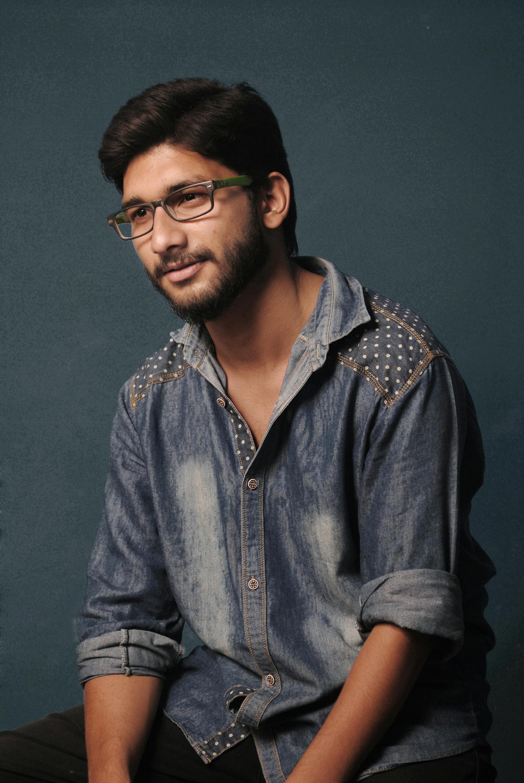 Casual studio portrait of a bearded man wearing glasses and a denim shirt against a plain background.