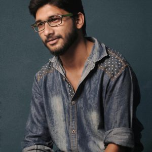 Casual studio portrait of a bearded man wearing glasses and a denim shirt against a plain background.